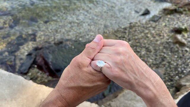 Elegant oval diamond ring showcased outdoors on the Croatian coast as loving couple hod hands with sparkling water and scenic background.