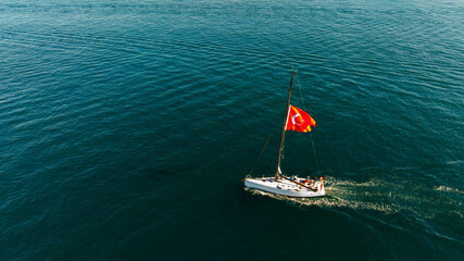 Aerial view Sailboat With Turkish Flag Cruising The Open Sea Under Clear Sky. A lone sailboat glides across calm blue waters, its bright Turkish flag fluttering from the mast. The scene conveys travel