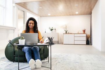 A woman with curly hair smiles while working on her laptop in a comfortable living room. She is seated on a bean bag chair, surrounded by plants and light decor.