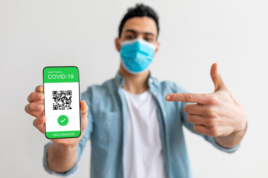 A young individual stands against a plain background, pointing at their smartphone, which shows a digital COVID-19 vaccination passport. The person wears a protective face mask and appears happy.