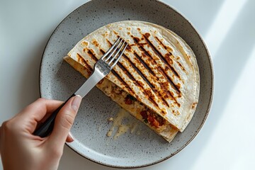 A person's hand holds a fork over a delicious, grilled quesadilla on a speckled grey plate, showcasing the crispy tortilla and savory filling, ready to be enjoyed.