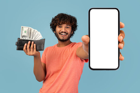 A cheerful young eastern man stands against a blue background, proudly displaying a blank smartphone and a wallet overflowing with cash.