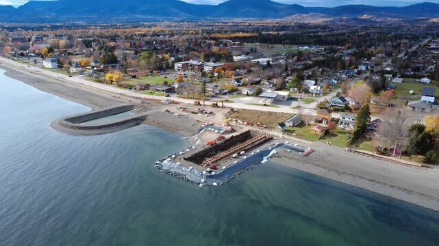 A coastal town is building temporary seawalls for beach replenishment work. Autumn trees line the streets and mountains rise in the distance, highlighting the development of the seaside community.