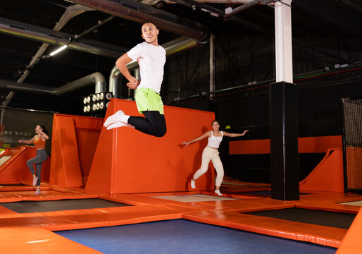 Cheerful young man in sportswear enjoying high jump, surrounded by bright colorful setting of modern indoor trampoline park ..