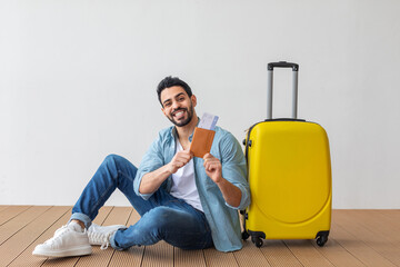A young man sits on a wooden floor with a beaming smile, holding his passport and travel tickets. Next to him, a bright yellow suitcase awaits his journey, symbolizing excitement and readiness.