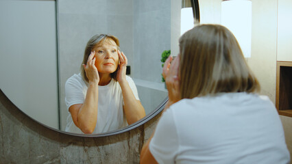 Elderly woman applying skincare in front of a mirror in a well-lit bathroom while enjoying a self-care moment