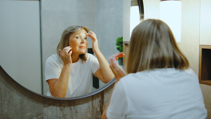 Elderly woman applying skincare products while looking in the mirror in a modern bathroom during the day