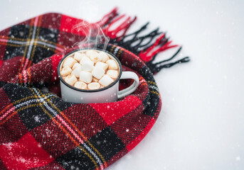 Cozy winter scene: hot chocolate with marshmallows in a warm scarf. A steaming mug of hot chocolate topped with marshmallows sits nestled in a red plaid scarf, perfect for a cold day
