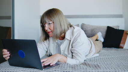 Elderly woman working on laptop while lying on bed at home during daytime, enjoying computer use and relaxation in a cozy setting