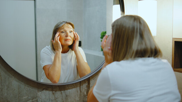 Elderly woman applying skincare in front of a mirror in a well-lit bathroom while enjoying a self-care moment