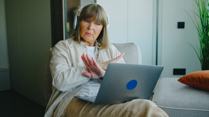 Elderly woman wearing headphones gestures while engaging with a laptop in a modern living room setting during the day