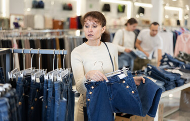 Among large selection of trousers on rails in store, woman stopped near showcase with jeans, holds...