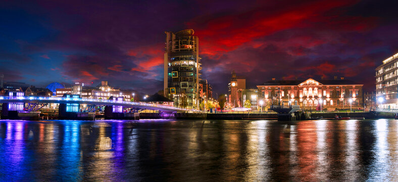 Belfast City night skyline with the Lagan Weir Pedestrian Bridge and colorful reflections on the River Lagan in Northern Ireland, United Kingdom, tranquil and vibrant green space riverwalk landscape.