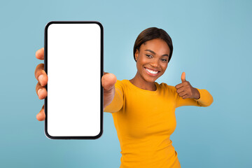 A young woman stands in front of a blue background, smiling widely as she holds up a smartphone...