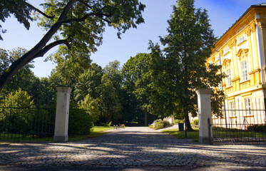 Fototapeta premium Entrance gate to the castle park and with the castle building on the right side. Castle building with yellow facade. Monuments of the Czech Republic. Traveling on summer holidays.