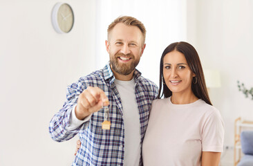 Portrait of happy young family couple standing indoors and holding keys from their new home. Smiling husband and wife celebrating buying family real estate and beginning of new chapter in their lives.