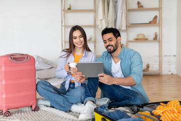 A couple sits on the floor in a bright and cozy room, looking at a tablet together while surrounded by luggage and clothing. They appear excited about their travel plans.