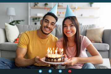 A man and woman, both wearing party hats, smile at the camera while holding a decorated cake with candles. They are celebrating a birthday remotely. The cozy room is decorated for the occasion.