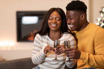 Two happy African American spouses laugh together while holding glasses of wine. They are celebrating either Valentine's Day or New Year indoors, sharing joyful moments by a cozy fireplace.
