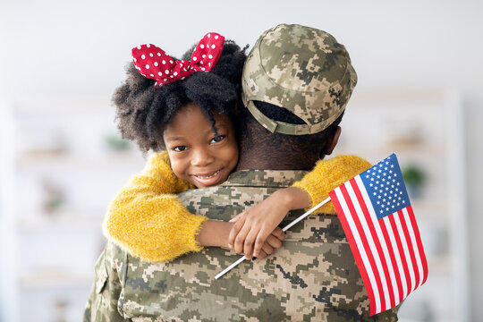 A joyful moment as a young girl embraces her soldier parent, holding a small American flag. The room feels warm and welcoming, celebrating their reunion.