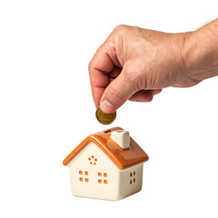 A hand places a coin into a ceramic house-shaped piggy bank. The bank has a brown roof and white walls with small windows. The background is transparent.