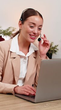 Young businesswoman at home office wears headset answers support call greets caller and asks details. Freelancer girl at table listens with care confirms ticket gives answer and keeps friendly tone