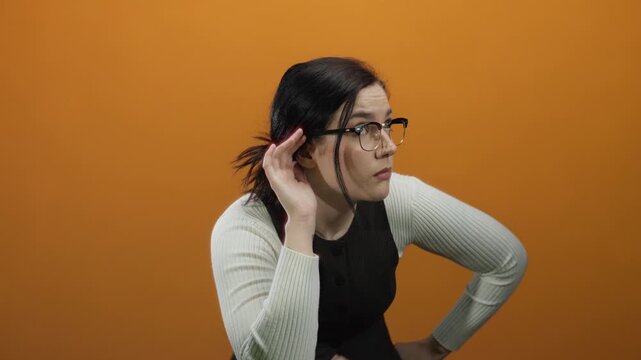 Woman with glasses and brunette hair gestures in front of an orange background, conveying curiosity and attentiveness.