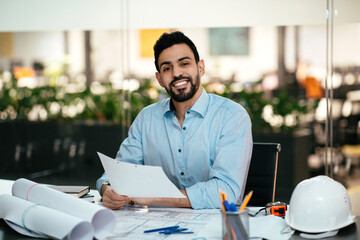 A young man sits at a desk in a contemporary office, smiling while holding architectural plans. The workspace is well-lit with greenery in the background.