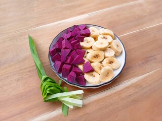 A plate filled with pieces of banana and purple sweet potato with pandan leaves on a wooden background. Healthy meal. Ingredients for healthy food