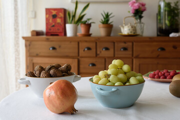 Primer plano de una mesa con alimentos saludables: cuencos de nueces y uvas, granada, kiwis, frambuesas.. y al fondo desenfocado cómoda de madera con macetas y adornos en su superficie. Vista frontal.