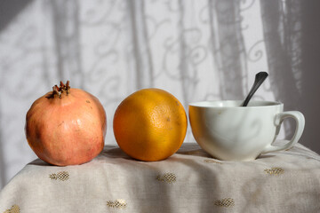 Bodegón formado por granada, naranja y taza blanca con cuchara en su interior. Vista frontal. Luz lateral con proyección de luces y sombras en los objetos y el fondo.