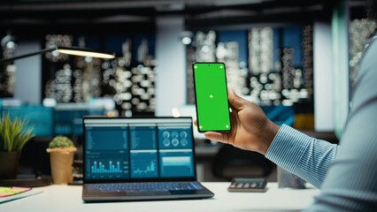 Black professional looks at chroma key on mobile phone screen at desk, solving business management tasks after hours. Male staff oversees project development, company objectives.