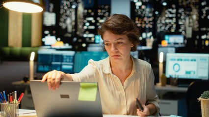 Female employee checking the time on her phone and closing laptop, preparing to leave office late...