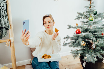 Happy Young Woman in cosy sweater taking selfie photo while relaxing on floor cusions near potted...
