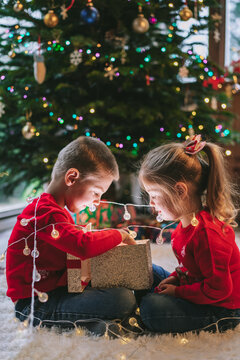 Two smiling kids in festive sweaters sitting under Christmas tree at home. Emotional sister and brother feeling excited of unwrapping Christmas gifts, having fun, childhood miracle time concept.