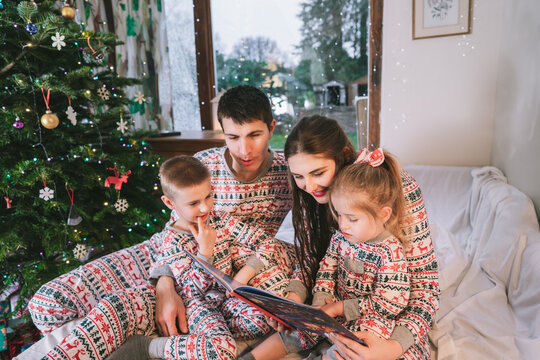 Happy family in festive pajamas reading book in bed in Christmas Eve. Happy parents and kids cuddling. Family time together. Happy childhood. Family celebrating winter holidays at decorated home.