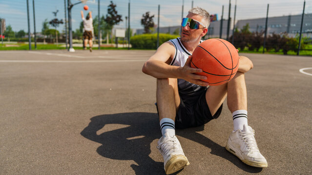 Casual basketball player sits on court, holding ball while teammate practices in background