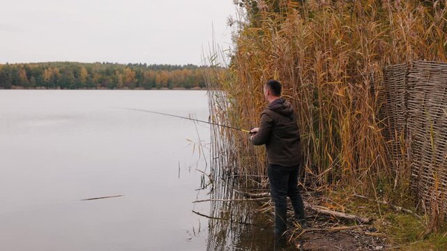 Fisherman casting spinning rod while predator fishing, man standing near reeds on autumn lake, outdoor sport fishing action and nature recreation concept with calm water and forest reflection