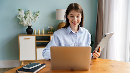 Young woman works on laptop while holding tablet at home desk