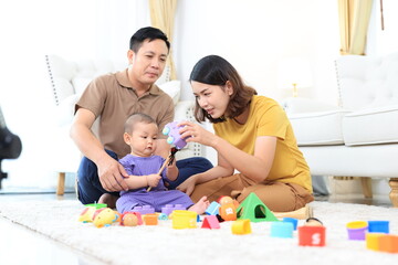An asian family of three, a woman and two men, are playing with colorful toys on the floor. Asian baby boy is holding a toy and the parents are clapping. Scene is happy and playful