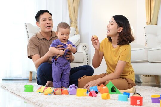 An asian family of three, a woman and two men, are playing with colorful toys on the floor. Asian baby boy is holding a toy and the parents are clapping. Scene is happy and playful