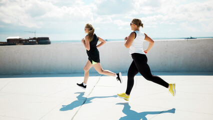 Two women jogging on a sunny rooftop with a city skyline in the background while wearing athletic...