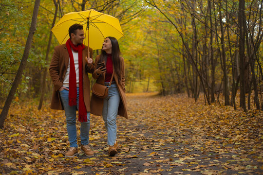 Smiling young couple walking under a yellow umbrella in an autumn park, dressed in brown coats and red accents, enjoying a romantic stroll surrounded by colorful fall leaves.