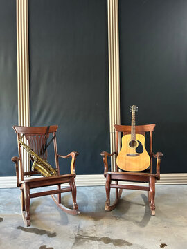 A guitar and a saxophone, both placed on two rocking chairs, on a cement floor, the wall black with reliefs in the shape of brown columns