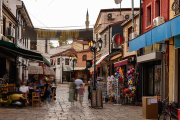 Naklejka premium Summer views of Old Bazaar in Skopje, North Macedonia, showing lively pedestrian streets with shops, cafes, market stalls, and mosque minaret rising above rooftops in background.