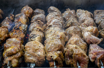 Multiple skewers of meat are grilling over a charcoal fire. The meat appears to be marinating and some pieces are already browning while wisps of smoke rise from the grill