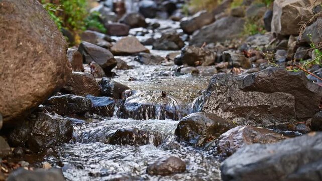 A stream of water flows between large rocks. The water is clear and the rocks are grey. The scene is peaceful and serene