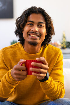 Indian male sitting at home holding red mug wearing mustard sweater and smartwatch by string lights