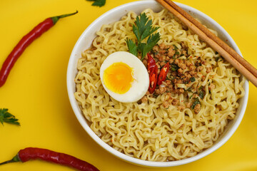 rA bowl of instant noodles topped with half-boiled egg, minced meat, and chili garnish on a bright yellow background.