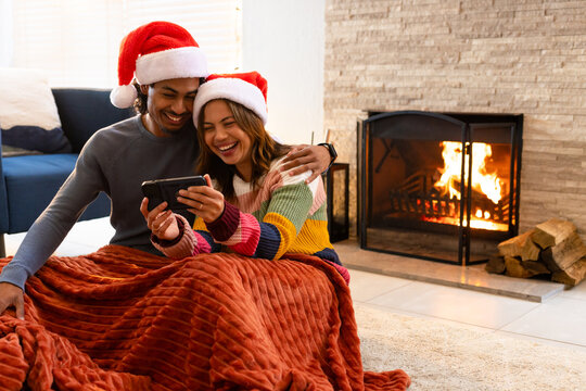 Couple huddling under blanket on living room floor by fireplace wearing Santa hats with smartphone